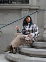 Mulher jovem com pele negra e cabelos escuros. Ela usa boné, camisa xadrez, calça e bota de cano curto e bico fino