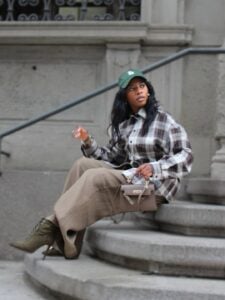 Mulher jovem com pele negra e cabelos escuros. Ela usa boné, camisa xadrez, calça e bota de cano curto e bico fino