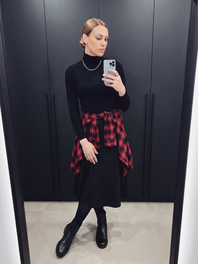 Mulher jovem, magra e de pele clara. Ela usa vestido preto midi de inverno, camisa xadrez amarrada na cintura e bota preta de cano curto