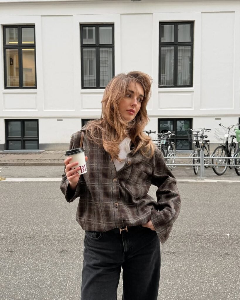 Mulher jovem, magra e de pele clara com cabelos loiros. Ela usa camisa xadrez, calça jeans e cinto
