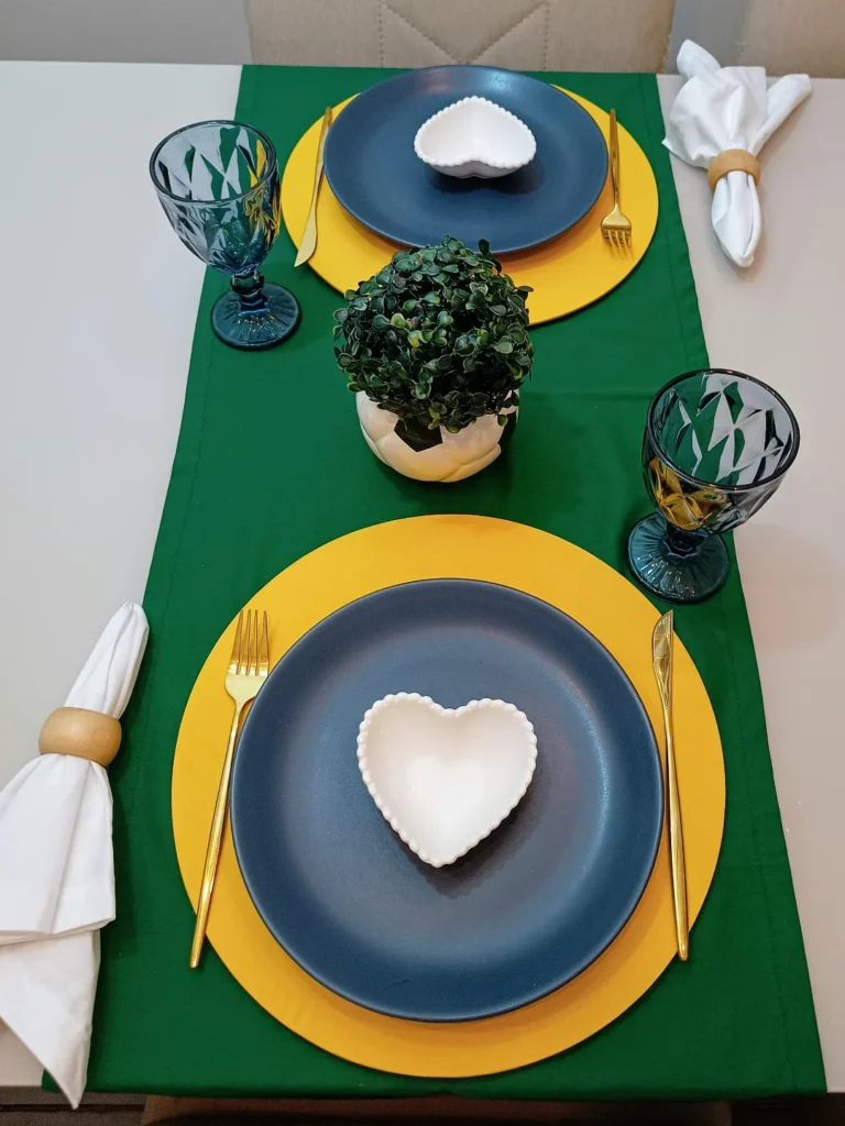 Mesa posta com caminho de mesa verde, sousplats amarelos, pratos azuis e pratinhos brancos em formato de cora