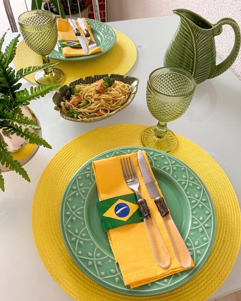 Mesa decorada para Copa com sousplats amarelos, pratos, taças e jarra verde, guardanapos amarelos com bandeirinha do Brasil