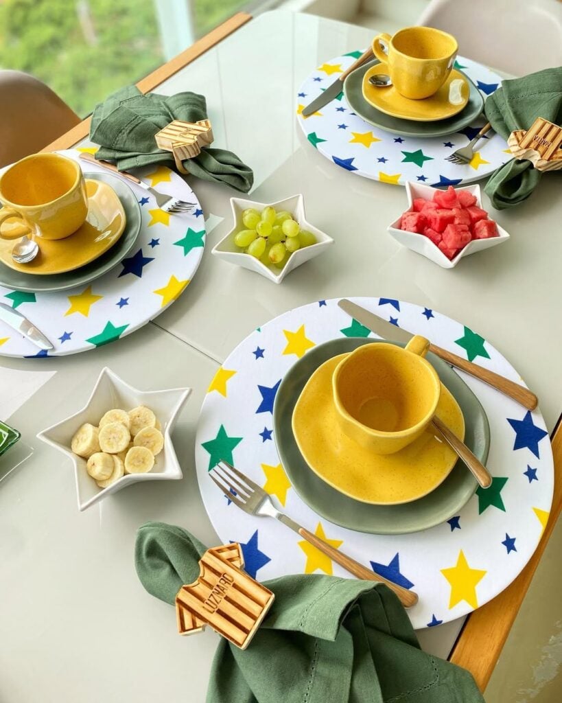 Mesa posta pra Copa com sousplats brancos com estrelas em verde, amarelo e azul, xícaras e pires em amarelo