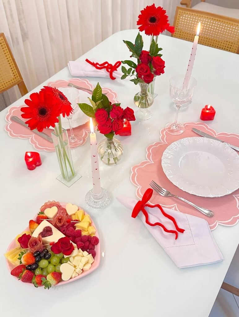 Mesa branca posta de Dia dos Namorados com jogo americano rosa, pratos brancos, e vasos transparentes com flores vermelhas