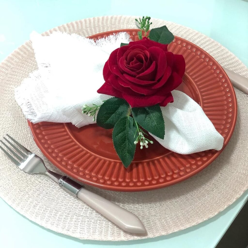 Mesa posta com sousplat rosa claro, prato em tom de terracota e guardanapo branco com anel em formato de rosa