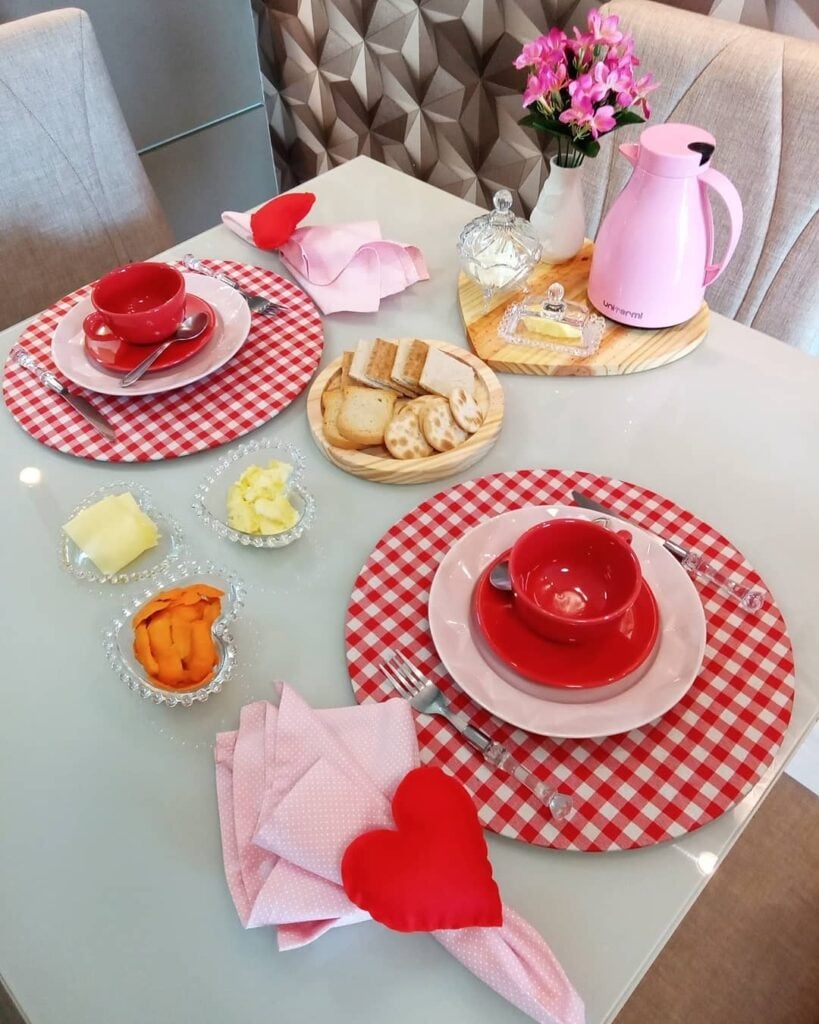 Mesa posta rosa e vermelho com sousplats xadrez, xícaras e pires vermelhos e guardanapos rosa com corações vermelhos