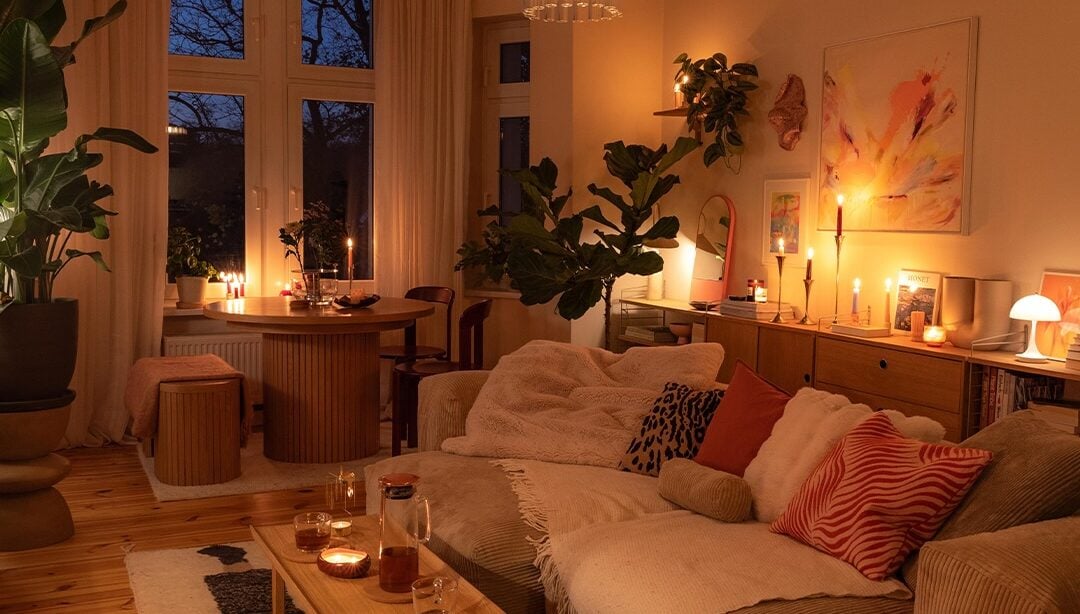 Sala com sofá em tom de bege, manta e almofadas, tapete grande e velas