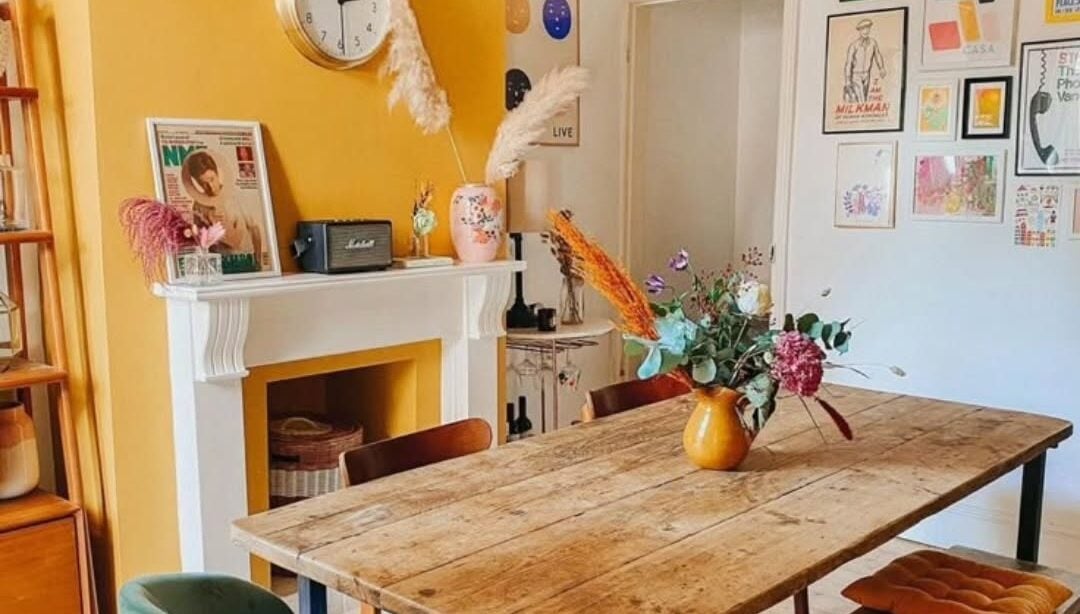 Sala de jantar com mesa e banco de madeira, cadeiras ao redor e parede amarela junto à lareira