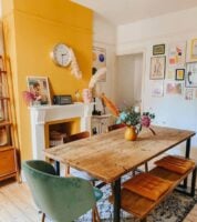Sala de jantar com mesa e banco de madeira, cadeiras ao redor e parede amarela junto à lareira