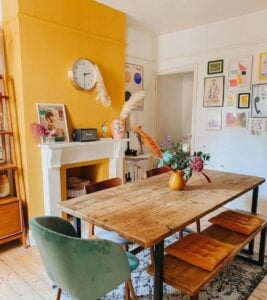 Sala de jantar com mesa e banco de madeira, cadeiras ao redor e parede amarela junto à lareira