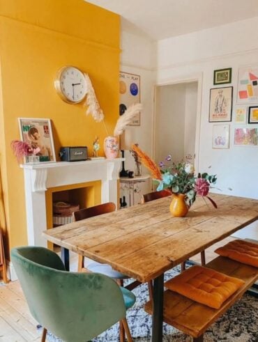 Sala de jantar com mesa e banco de madeira, cadeiras ao redor e parede amarela junto à lareira