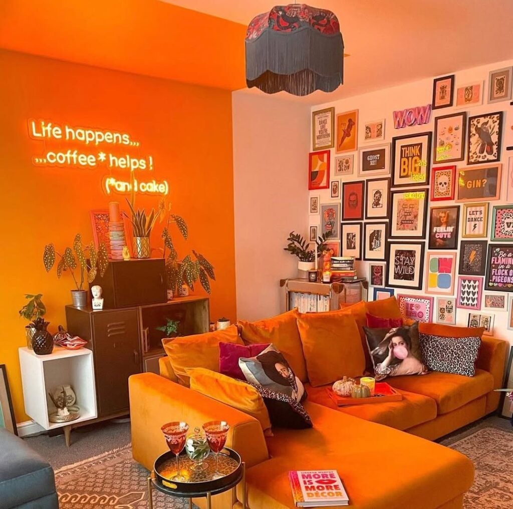 Sala com sofá e parede laranja com letreiro e parede branca com diversos quadros
