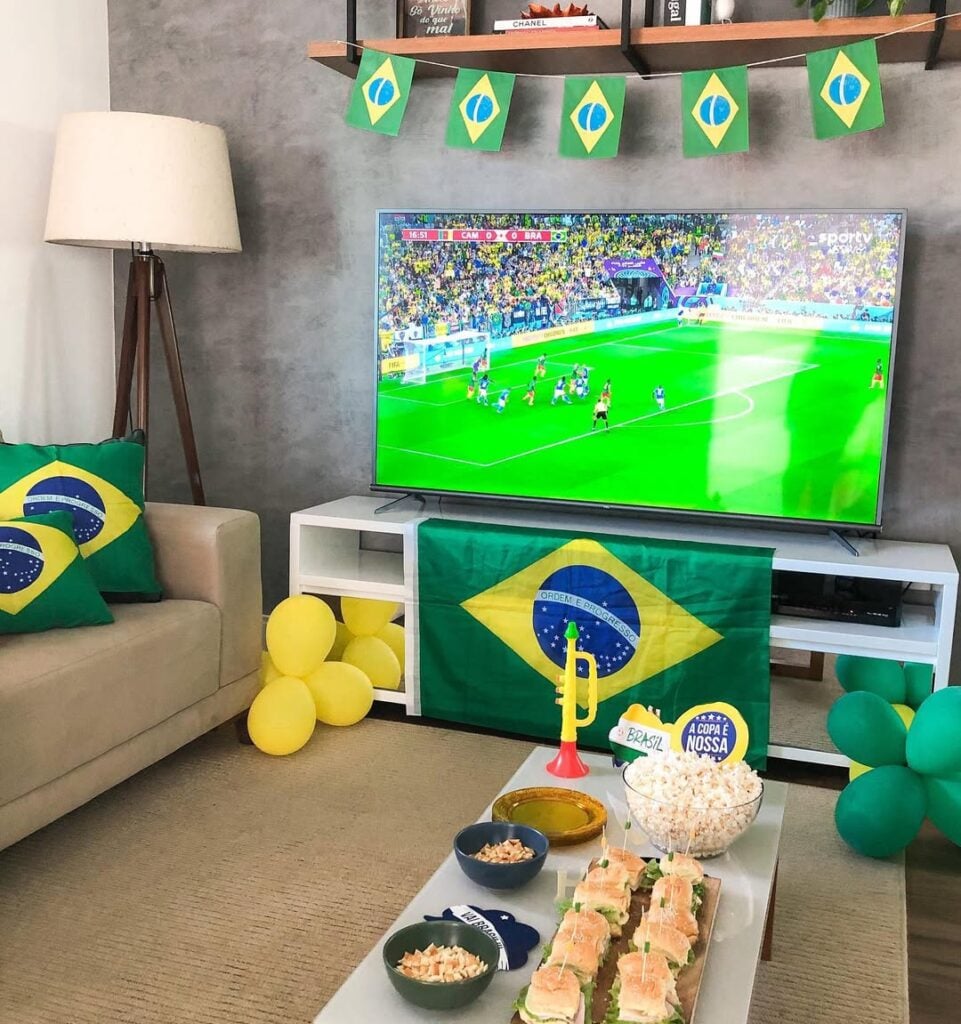Sala com rack branco, sofá neutro, bandeirinhas do Brasil e balões em verde e amarelo