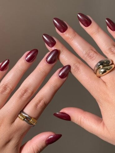 Mãos de pele clara, com anéis em dourado e prata e unha amendoada vermelho-cereja