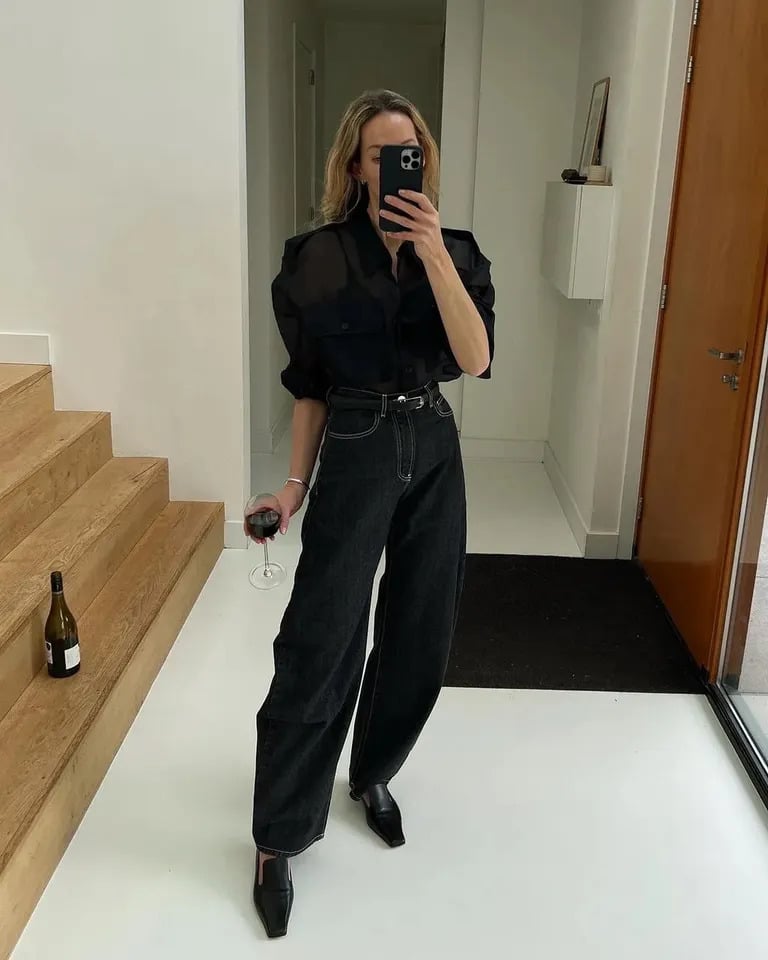 Mulher jovem, magra e de pele clara com look all black composto de camisa com manga bufante, calça jeans e mocassim