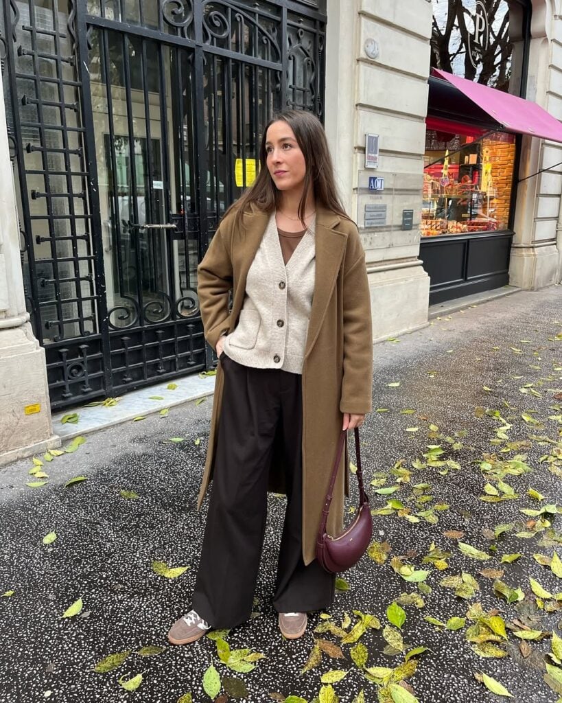 Mulher jovem, magra e de pele clara com cabelo castanho. Ela usa sobretudo marrom claro, suéter cinza claro, calça marrom escuro, tênis marrom claro e bolsa vermelho vinho