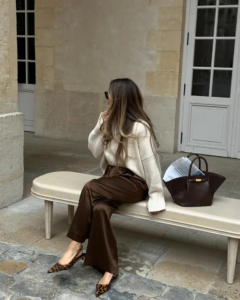 Mulher jovem, magra e de pele clara. Ela usa suéter off-white, calça de couro marrom e slingback animal print