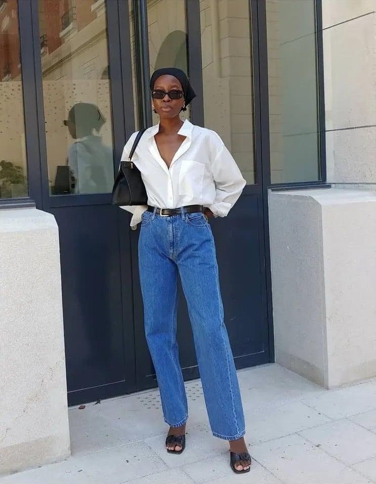 Mulher jovem, magra e de pele negra. Ela usa camisa branca, calça jeans com cinto, tamanco preto, óculos de sol, bolsa preta e bandana preta