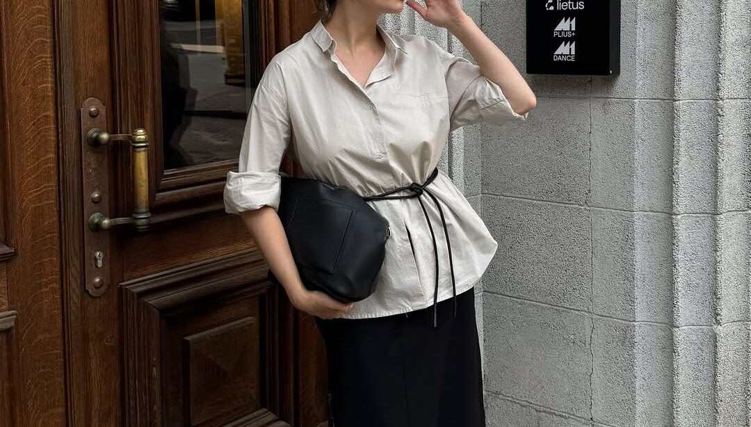 Mulher jovem de pele clara. Ela usa camisa, cinto de amarração, saia longa e chinelo