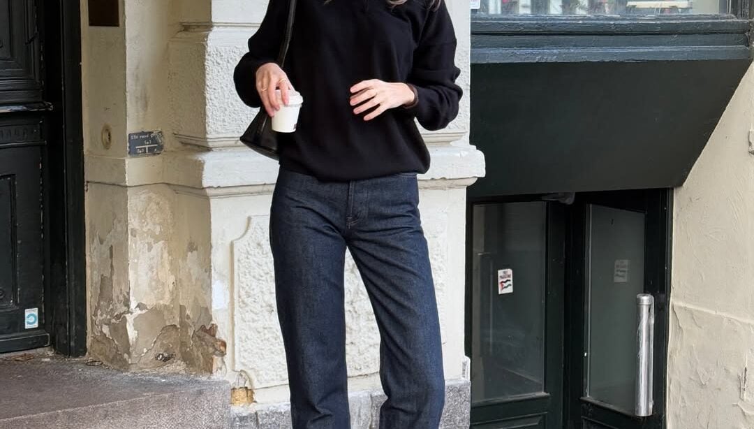 Mulher jovem, magra e de pele clara. Ela usa suéter preto, calça jeans com lavagem escura e bota preta de bico fino com salto alto
