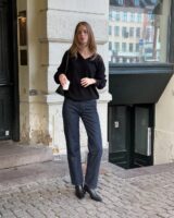 Mulher jovem, magra e de pele clara. Ela usa suéter preto, calça jeans com lavagem escura e bota preta de bico fino com salto alto