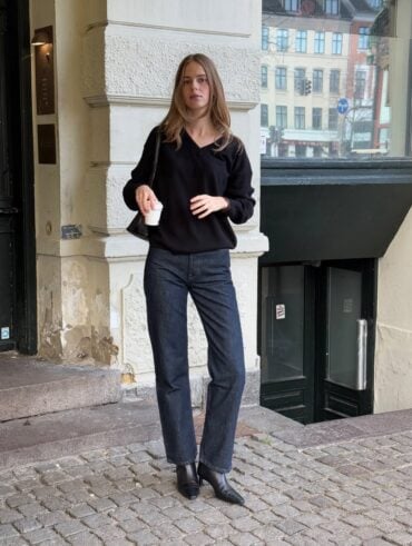 Mulher jovem, magra e de pele clara. Ela usa suéter preto, calça jeans com lavagem escura e bota preta de bico fino com salto alto