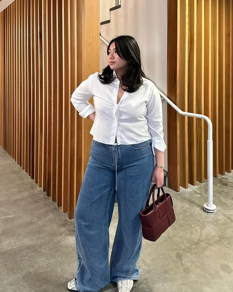 Mulher jovem mid size com pele clara e cabelos escuros. Ela usa camisa branca curta, calça jeans e mocassim estampado