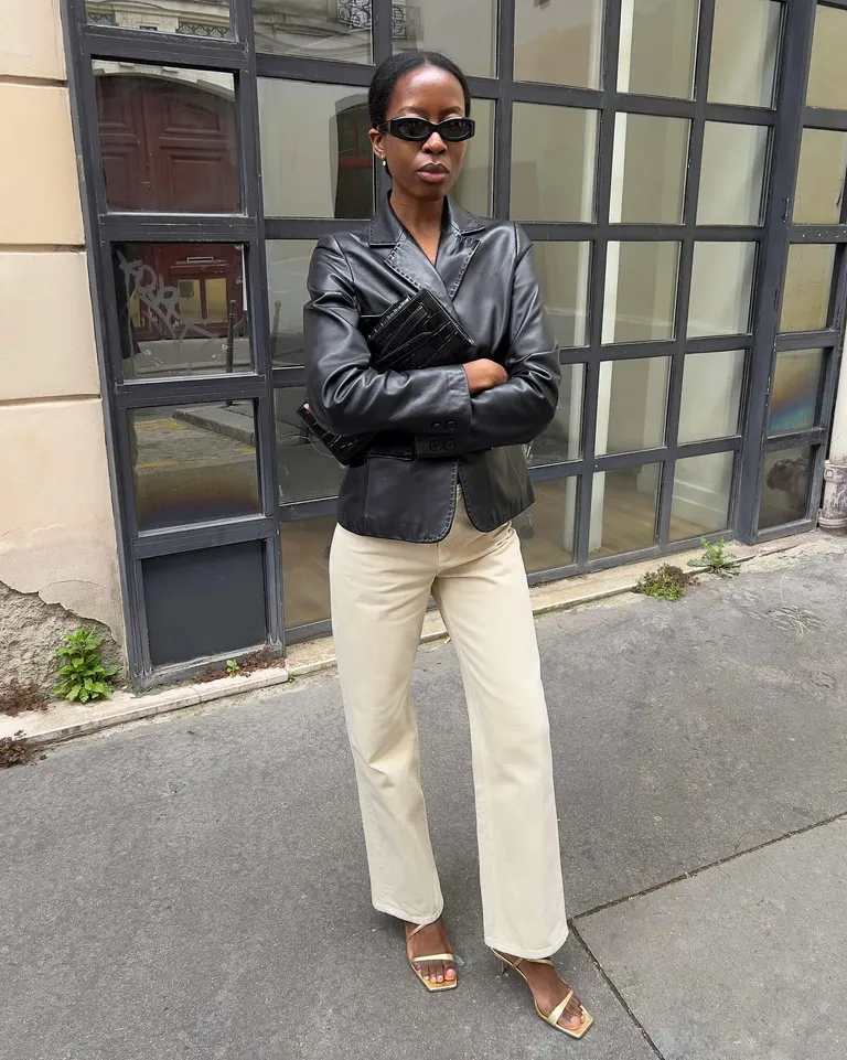Mulher jovem e magra de pele clara. Ela usa jaqueta de couro preta, calça jeans e salto alto