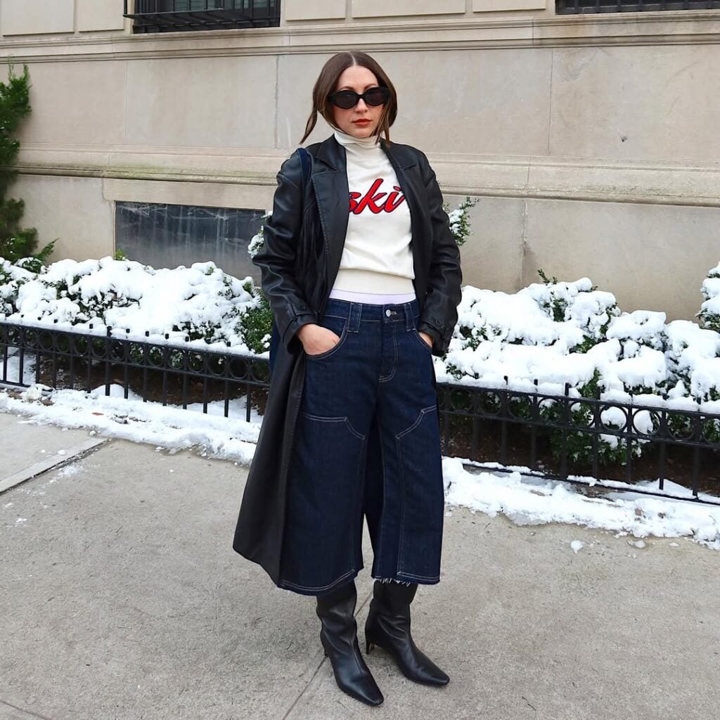 Mulher jovem, magra e de pele clara com cabelo castanho. Ela usa look de inverno com bota de cano alto