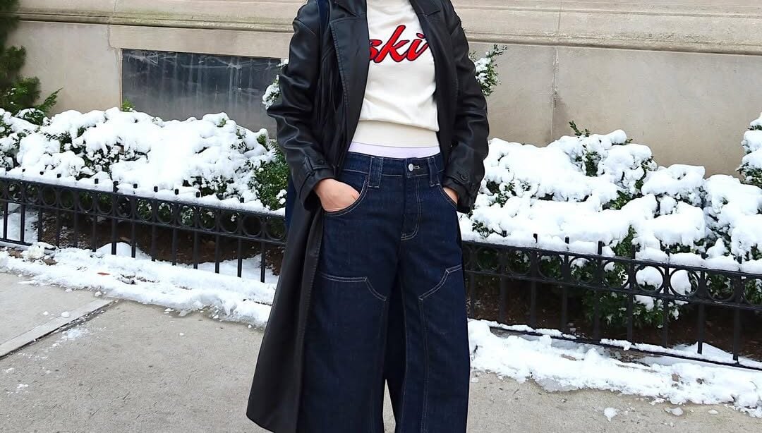 Mulher jovem, magra e de pele clara com cabelo castanho. Ela usa look de inverno com bota de cano alto