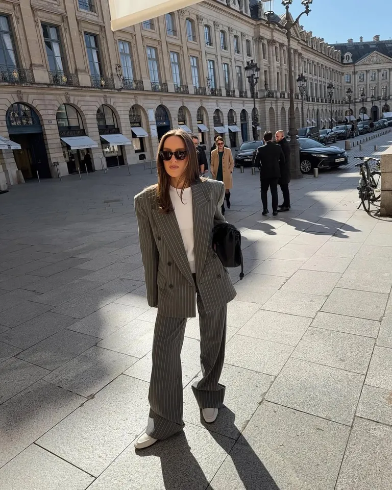 Mulher jovem de pele clara e cabelo castanho. Ela usa blazer, calça de alfaiataria e mocassim branco