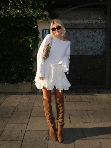Mulher jovem, magra e de pele clara com cabelos loiros. Ela usa vestido branco e bota de cano longo marrom