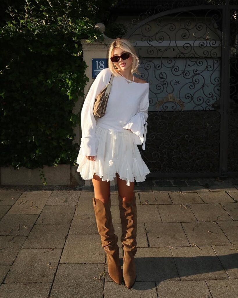 Mulher jovem, magra e de pele clara com cabelos loiros. Ela usa vestido branco e bota de cano longo marrom