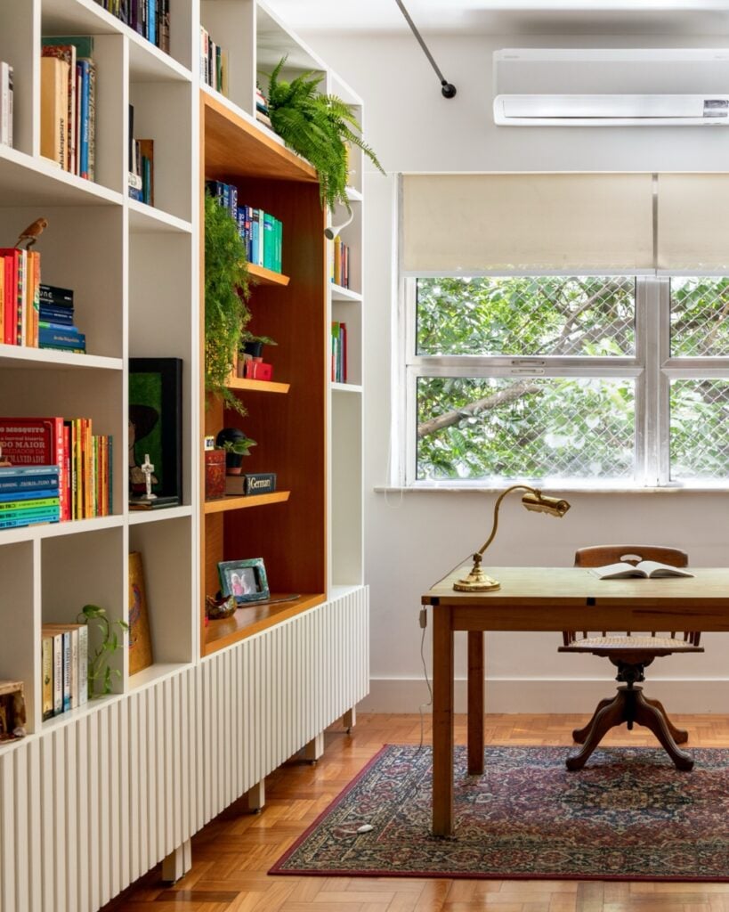 Ambiente com estante branca, livros, objetos decorativos e mesa de madeira com cadeira e luminária
