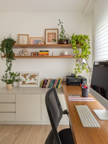 Home office com bancada de madeira, computador, impressora e balcão lateral com livros, quadro e planta