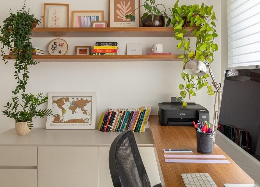 Home office com bancada de madeira, computador, impressora e balcão lateral com livros, quadro e planta