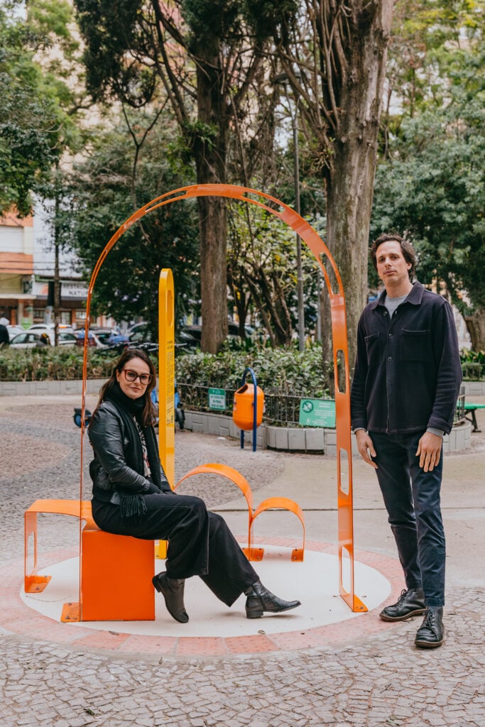 Mulher vestida de preto sentada em estrutra de metal laranja, homem em pé com roupa escura