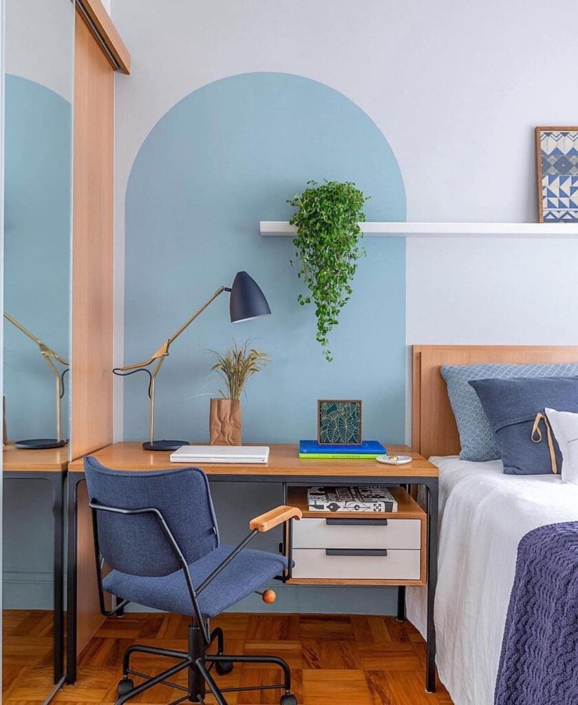 Quarto azul com pintura arredondada atrás da escrivaninha, prateleira branca com quadros e planta
