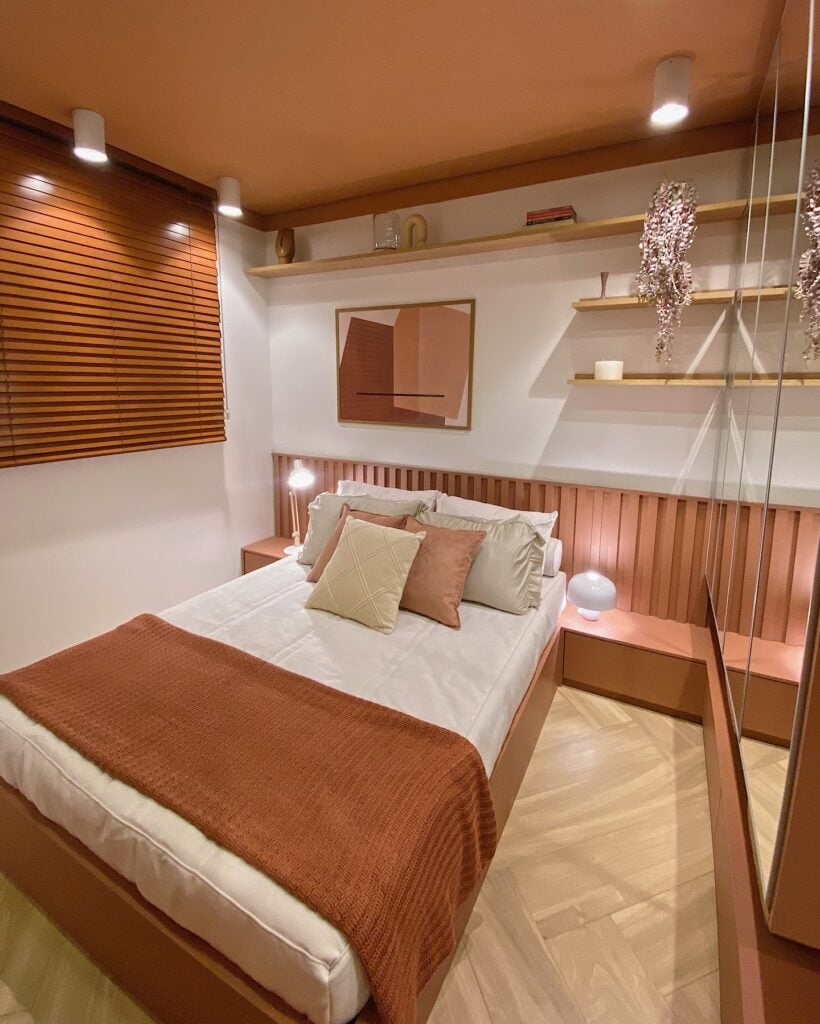 Quarto com paredes brancas e tons terrosos na cabeceira, cortina e roupas na cama