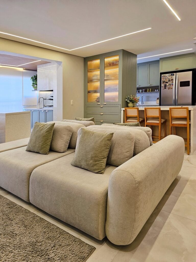 Sala com sofá confortável em tom de cinza e ao fundo a cozinha com armários verde