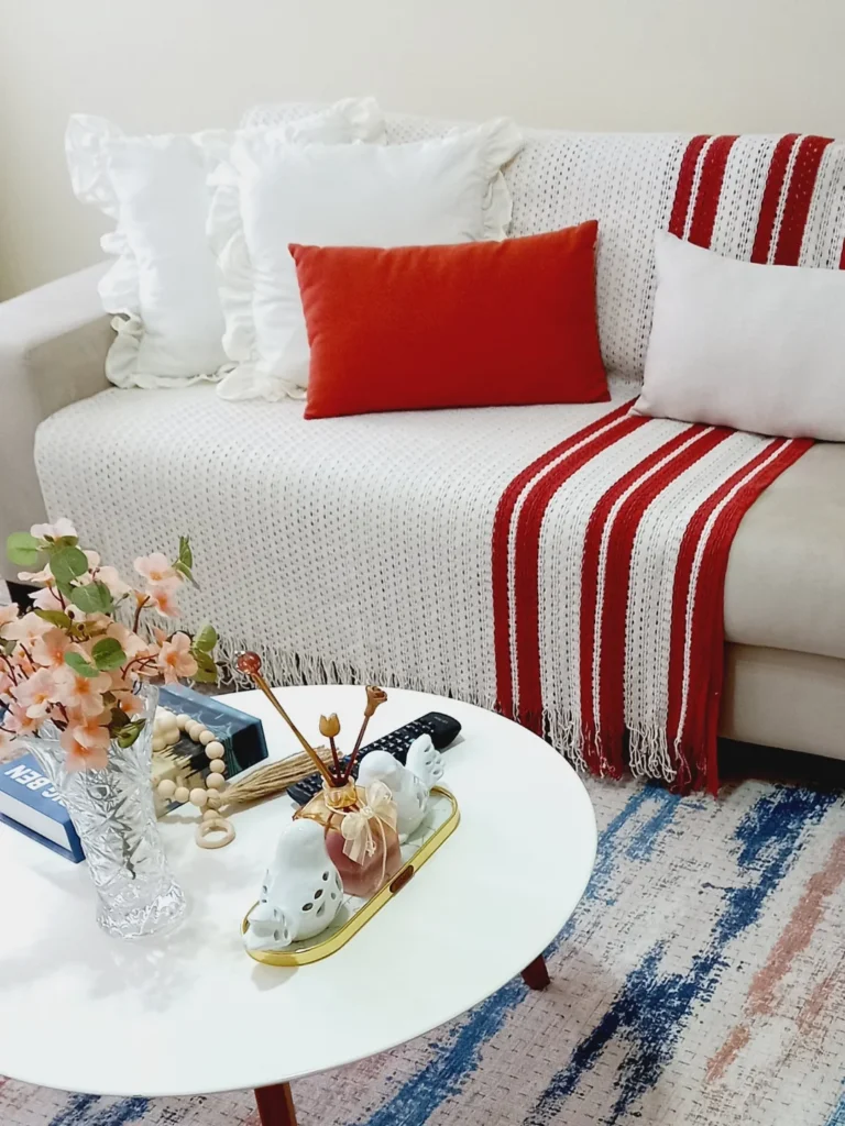 Sofá claro com almofadas e manta em branco e vermelho, mesa de centro branca com objetos decorativos