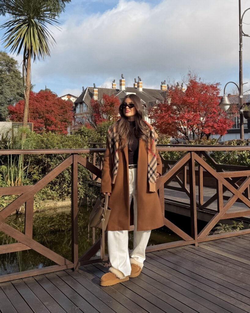 Mulher jovem e magra com cabelo castanho esucuro. Ela usa casaco longo, calça, blusa e bota ugg