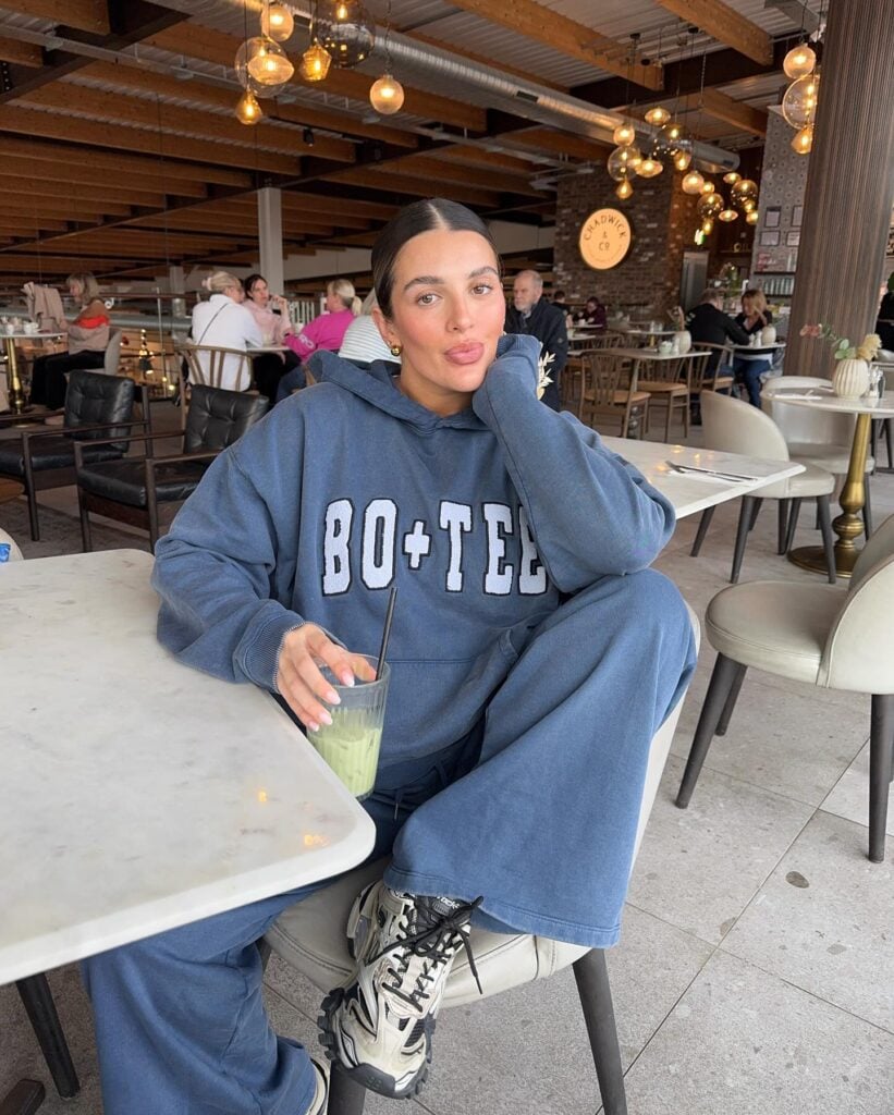 Mulher jovem e magra de pele clara. Ela usa conjunto de moletom azul e tênis esportivo