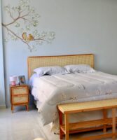 Quarto com parede azul-claro, cabeceira, mesa de cabeceira e banco de palhinha e pintura com passarinhos na parede