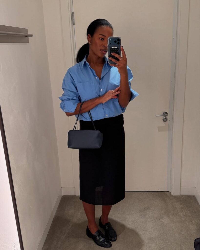 Mulher jovem de pele negra com camisa azul, saia preta e mocassim preto