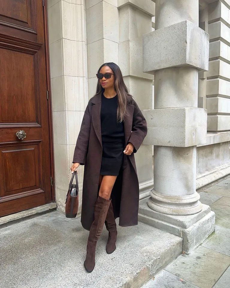 Mulher jovem e magra de pele negra. Ela usa vestido, sobretudo e bota de cano alto