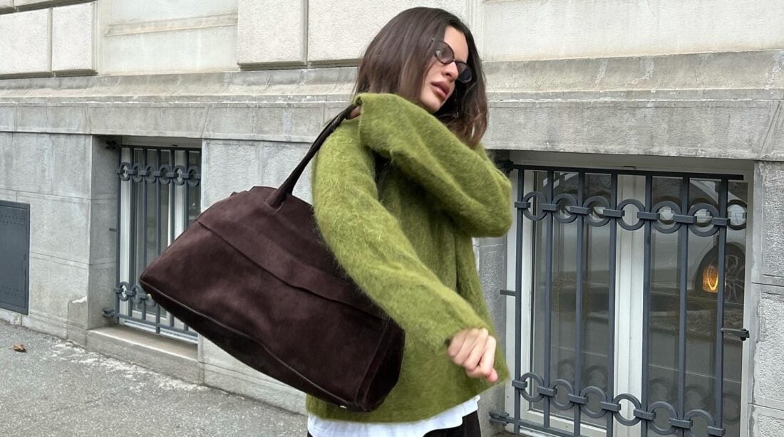 Mulher jovem e magra de pele clara e cabelo castanho. Ela usa suéter verde e calça preta