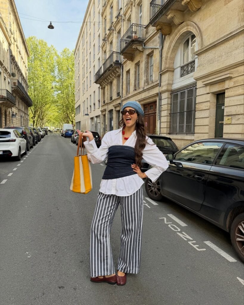 Mulher jovem, magra e de pele clara. Ela usa camisa branca, top jeans, calça listrada e sapatilha tabi