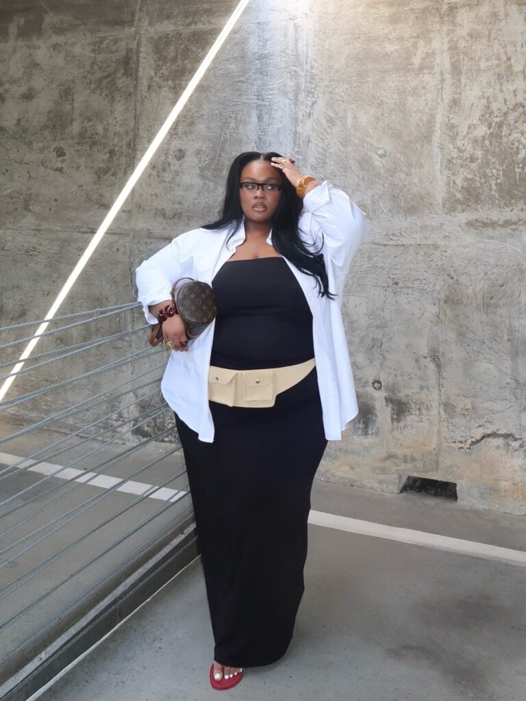 Mulher jovem, gorda e com cabelo escuro. Ela usa camisa branca, vestido preto longo e chinelo