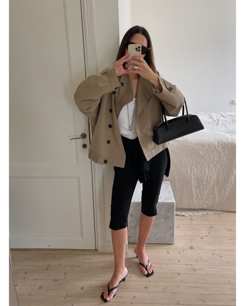 Mulher jovem, magra e de pele clara. Ela usa trench coat curto, blusa btanca, calça capri preta e chinelo kitten heels preto